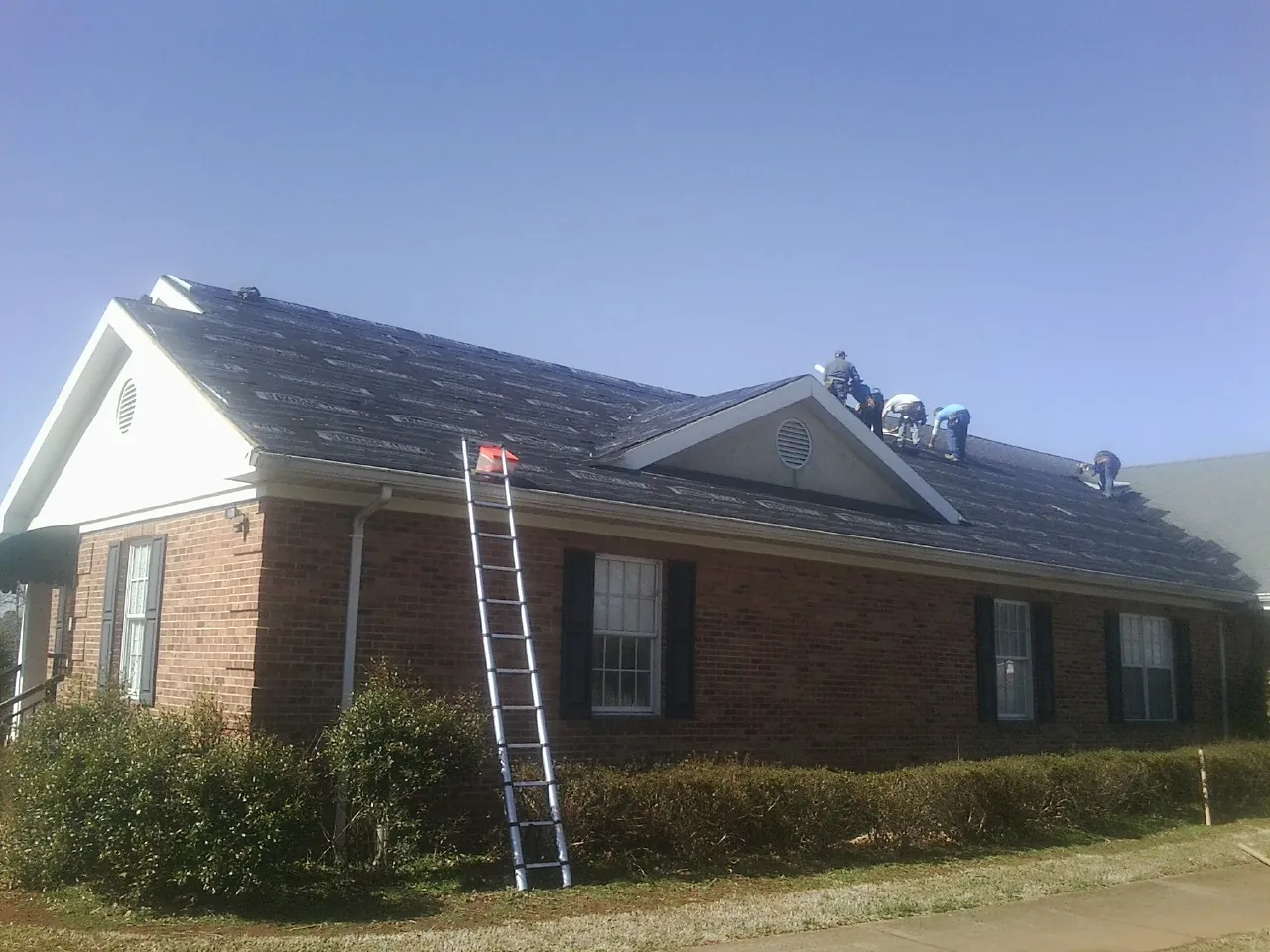 Crew tearing shingles off a brick ranch roof with a ladder set — Emergency Roof Repair work by our crew in Beloit