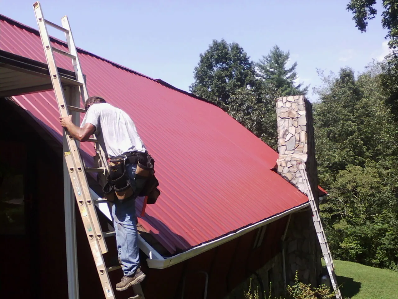 Roofer on a ladder beside a red metal roof and stone chimney — Soffit & Fascia Repair by Roofing Brothers in Beloit